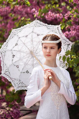 Young beautiful woman in white historical dress and headpiece standing with umbrella among trees with lilac flowers. Historic portrait of aristocratic lady