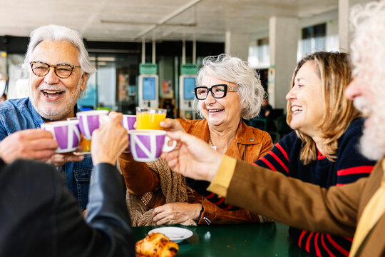 Happy group of senior people enjoying coffee at cafeteria bar. Retired generation community having fun gathered on terrace toasting hot drink. Elderly friendship lifestyle concept.