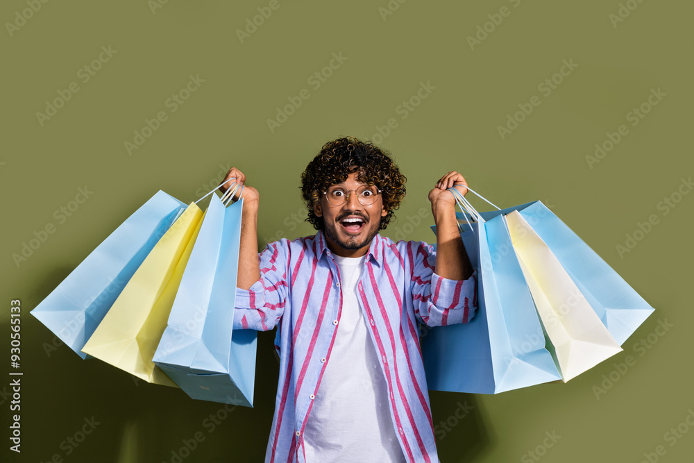 Sticker Photo of impressed guy with wavy hair dressed striped shirt in glasses raising up shopping bags isolated on khaki color background