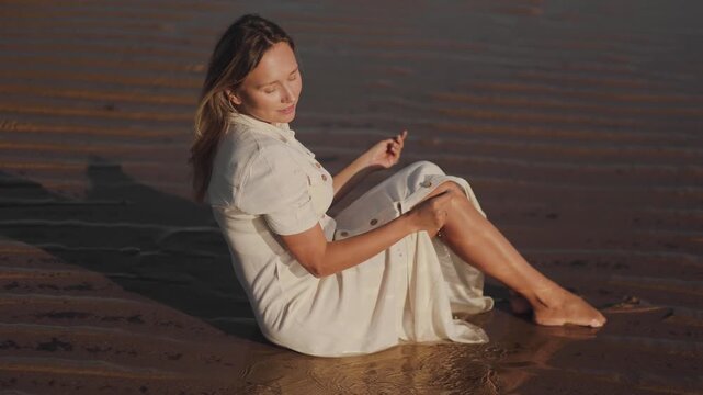 Cinematic shot of a luxurious blonde lying in the dunes, on the wet sand near the ocean, model, woman posing in a beige dress with wet hair, tanning, tourism, vacation, luxury, golden light, care  