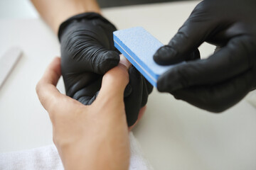 Close-up of gloved hands providing manicure service using a nail buffer block on client's fingernail, focusing on details of the process, and ensuring precision in beauty treatment