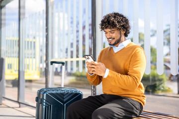 Smiling man sitting at bus stop using smartphone with suitcase beside him. Sunny day setting highlighting casual travel and connectivity. Ideal for travel, transportation, communication themes. © Liubomir