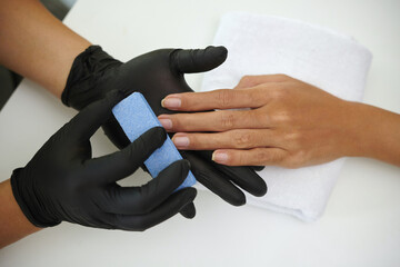 Hands of a spa professional in black gloves providing manicure on a client's hand using a blue nail file, set against a clean, white surface creating a serene environment
