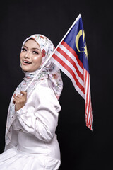 Naklejka premium Beautiful Asian girl wearing white gress with hijab, happily holding Malaysian flag isolated over black background. Independence day, new year nationalism concept.
