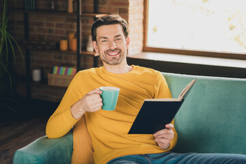 Photo portrait of young guy sit couch read book drink coffee dressed yellow garment spend pastime...