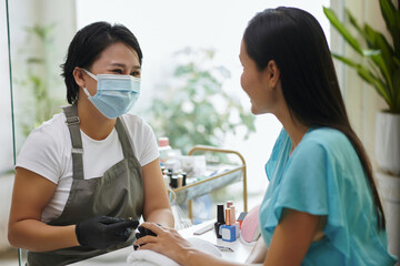 Beautician wearing mask and gloves while offering manicure service to client in well-lit salon environment. Both individuals engaged in conversation creating a friendly atmosphere