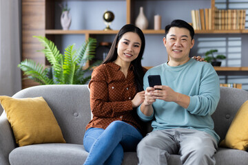 Cheerful Asian couple sitting on comfortable sofa at home, engaging with a smartphone. They appear relaxed and happy while sharing moments, highlighting themes of family, technology, and connection.