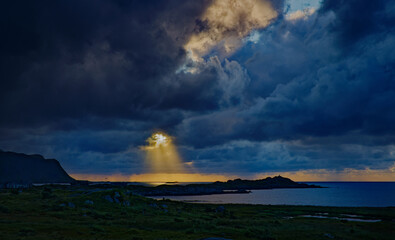 The sun breaks through storm clouds at sunset creating drama over the mountains and rocky coast. Lofoten Islands, Northern Norway.