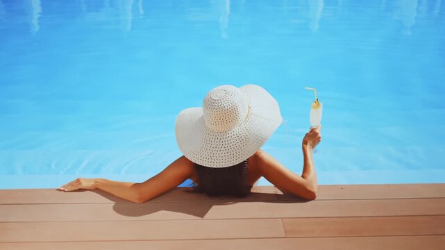 Woman in wide-brimmed hat relaxes at edge of pool, holding and refreshing drink. Concept of leisure, summer relaxation, and poolside luxury
