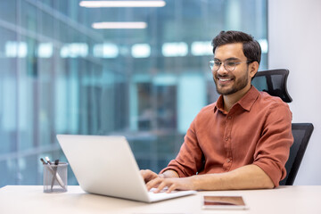 Confident man in casual attire working on laptop at office desk, with phone nearby. Bright, modern...