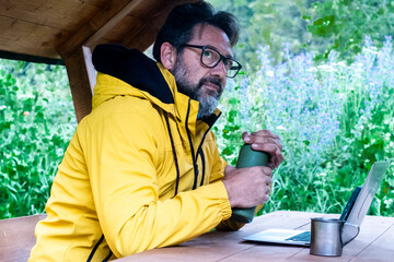 One man with yellow jacker opening metal bottle of coffee sitting ona wooden table at the mountain waiting to work on laptop with roaming data connection. Male mature people in outdoor leisure