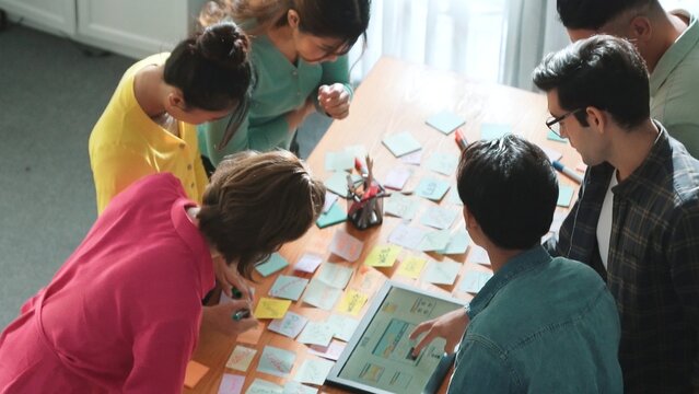 Top view of manager writing idea on sticky notes and explain idea about web design. Professional marketing team or programmer brainstorming idea while looking at tablet with color palette. Symposium.