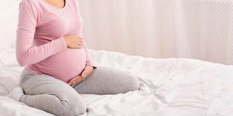 Pregnancy Concept. Unrecognizable Pregnant Lady Sitting On Bed Touching Belly In Bedroom. Cropped, Panorama With Free Space For Text