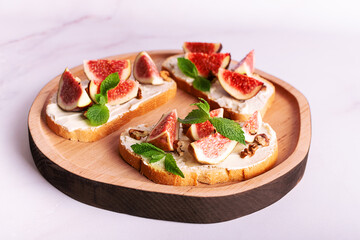 Tasty homemade sandwiches with sweet figs, cream cheese, walnuts. honey and mint leaves on the wooden serving plate on light marble background close up.