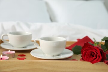 Wooden tray with burning candles, rose, cups of drink and red paper hearts on bed indoors