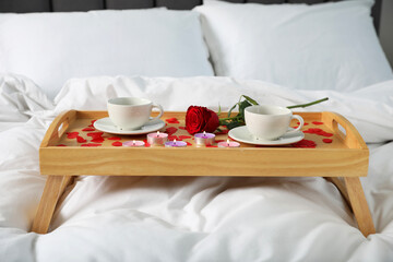 Wooden tray with burning candles, rose, cups of coffee and red paper hearts on bed indoors