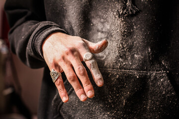 Dusty fingers of a woodworker, embellished with ornate rings, demonstrate the blend of artistry and labor in woodworking.