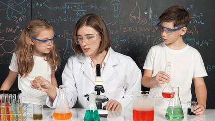 Caucasian boy mixing chemical liquid while teacher giving advice. Professional instructor wearing lab suit looking for diverse student at table with beaker filled with colored solution. Erudition.