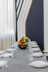 Elegant dining table setting with white plates, glasses, and a fruit centerpiece in a modern interior.
