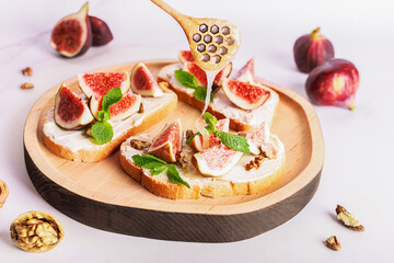 Tasty homemade sandwiches with sweet figs, cream cheese, walnuts. honey and mint leaves on the wooden serving plate on light marble background close up.