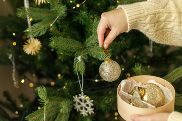Hands in knitted sweater holding stylish Christmas ornaments in wooden box on background of christmas tree with golden lights. Decorating christmas tree with vintage golden baubles