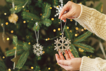 Hands in knitted sweater hanging stylish Christmas ornament on christmas tree with golden lights. Decorating christmas tree with vintage glass snowflake