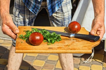one kitchen sharp dangerous black handle shiny new iron knife lies on cutting wooden board with two red fresh vitamin tomatoes and chopped green parsley in hands of man during daytime outdoors