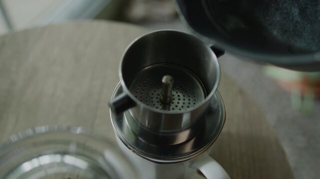 In a close-up shot, water is poured into a Vietnamese coffee phin, slowly dripping through the grounds to brew fresh coffee in the morning, with lush plants softly blurred in the background.