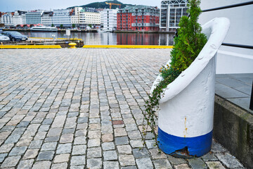 Waterfront scene in Bergen, Norway, featuring cobblestone paving, urban architecture, and a decorative planter made from a repurposed marine bollard