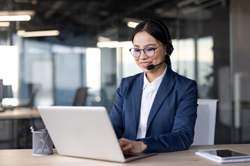 Asian businesswoman with headset using laptop in modern office. Professional and engaged in task with focus on technology.