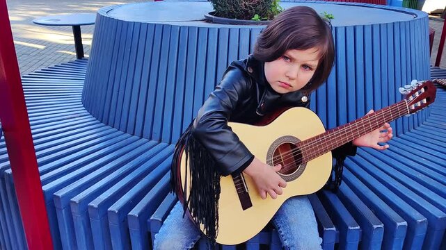 A small, beautiful girl in jeans and a leather jacket plays a six-string acoustic guitar, alone on the street