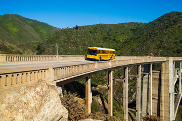 A yellow school bus crossing a historic Bixby bridge surrounded by lush green hills under a clear blue sky