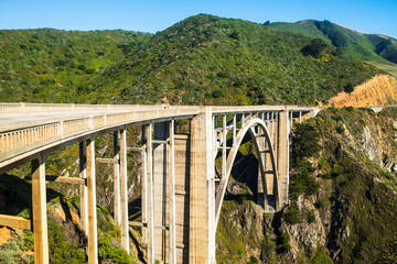 crossing a historic Bixby bridge surrounded by lush green hills under a clear blue sky