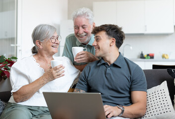 Obraz premium Happy multigenerational family sitting relaxing on sofa, grandparents drink a coffee cup while young grandson uses laptop. Positive emotions