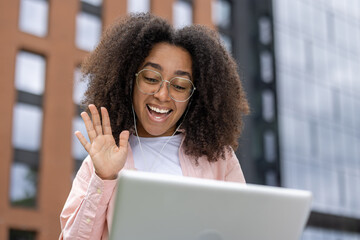 Smiling young woman engaged in a video call, waving at her screen while sitting outside. Enjoying modern technology to stay connected with loved ones.