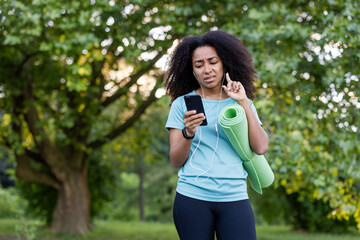 A woman in a park listens to music while checking her phone, holding a yoga mat, with a concerned expression.