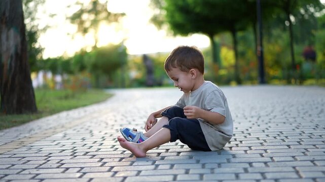 Cute little toddler boy sits on the wide paved alley. Kid takes off his shoe and throws in the direction of camera. Blurred backdrop.
