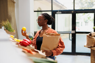 Young black woman holding fresh lemon and brown paper bag in eco friendly bio food shop. African american female customer examining freshly harvested produce in local convenience store.