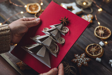 Handmade New Year origami postcard with paper christmas fir tree. Holiday idea for family time and gift for beloved. Gingerbread cookies, lights, wooden background. Zero waste, recyclable materials