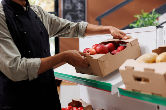 Store worker wearing black apron handles small crate of several red apples on a shelf in grocery store. Close-up of male vendor placing box of fresh fruits on white racks of eco friendly supermarket.