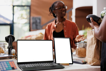 Black woman speaking with vendor at checkout counter while laptop and tablet display isolated white screens. Devices showing blank copy space mockup, ideal for advertising of eco friendly bio store.