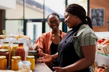 African American vendor selling fresh preserved chemical free products to consumer with spectacles. Smiling black women standing near glass jars conversing about healthy sustainable food recipes.