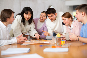 Cheerful emotional young Chinese female student playing board game at table with group of friends. Concept of socialization of young adults