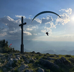 Gleitschirmflieger (Paraglider) im Abendlicht neben dem Gipfelkreuz der Hochries, Alpen, Chiemgau,...