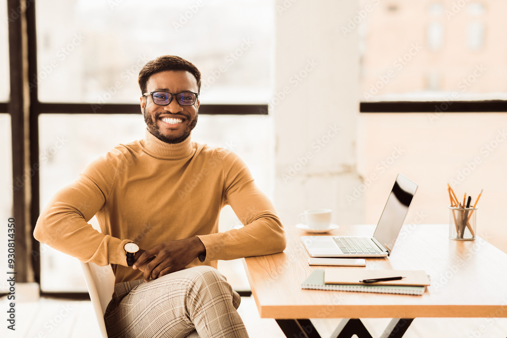 Poster enjoying work. smiling afro guy working on his laptop and looking at camera sitting at his working p