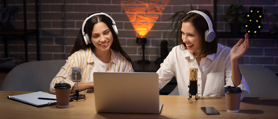 Young women with headphones and laptop recording podcast in studio at night