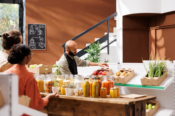 Middle Eastern storekeeper putting a box filled with red apples on shelf in eco friendly store. Male vendor arranges aisle of organic fresh products while two customers, male and female, look on.