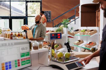 In sustainable grocery store, male customer purchases local grains and bulk products using plastic free bags. Middle eastern man pouring natural organic pantry food products from glass container.