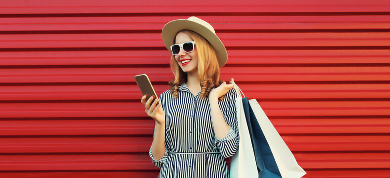 Beautiful happy young woman looking at phone with shopping bag in summer hat on red background