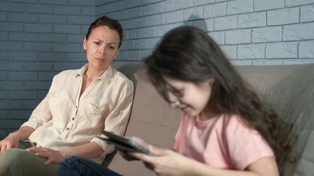 Mum look at daughter with online addiction indoor. A sad mother with problems with her daughter sitting by her during internet games.